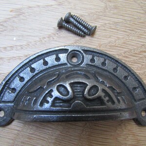 Op de afbeelding: Een donkergrijze gietijzeren komgreep met een sierlijk ontwerp en twee schroeven. De greep heeft een halfronde vorm. De schroeven zijn zilverkleurig en bevinden zich boven de greep.