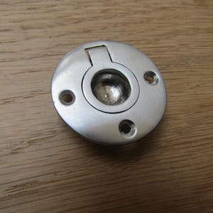 Puede incluir: Un tirador empotrado circular de metal cepillado con un centro empotrado y una lengüeta rectangular con bisagras. El tirador tiene tres pequeños orificios para tornillos y está diseñado para un montaje empotrado. El herraje es de color plateado.