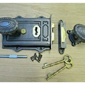 Könnte beinhalten: Ein antikes Türschloss-Set mit verzierten, oval geformten Griffen und einer rechteckigen Frontplatte. Das Set enthält ein Schließblech, zwei Schlüssel, Schrauben und eine Verbindungsstange. Die Metallkomponenten haben eine dunkle, gealterte Oberfläche.