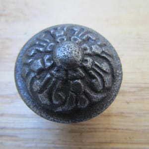 Könnte beinhalten: Runder Möbelknopf aus Metall im antiken Stil mit detailliertem floralem Design. Der Knopf hat eine dunkle, verwitterte graue Farbe und eine strukturierte Oberfläche. Dieses dekorative Hardware-Element eignet sich für Schränke und Schubladen, ca. 3 cm Durchmesser.