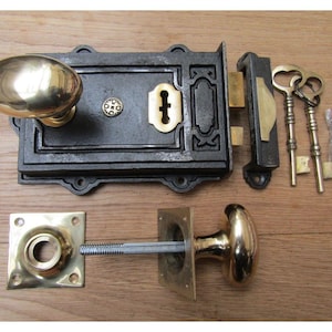 Può includere: Un set di serratura per porta vintage con una placca frontale in ghisa nera, una maniglia in ottone e due chiavi. Il set include una placca di chiusura in ottone e una maniglia in ottone abbinata. L'hardware è esposto su una superficie di legno.