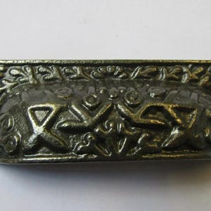May include: Antique bronze-colored drawer pull with ornate floral and geometric relief details. The rectangular pull has a curved handle and two mounting holes. A decorative hardware piece for furniture.