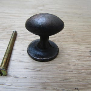 Könnte beinhalten: Ein dunkelbrauner, ovaler Schrankknauf mit kurzem Stiel und rundem Sockel. Eine goldfarbene Schraube befindet sich links vom Knauf. Die Hardware befindet sich auf einer hellen Holzmaserung.