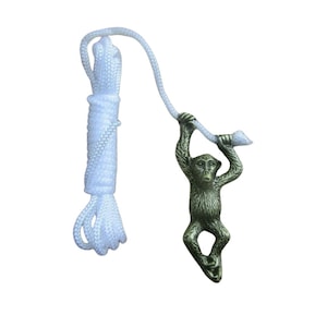 Op de afbeelding: Een bronzen aapfiguur hangt aan een wit touw. De aap is gedetailleerd met zichtbare vacht en gezichtskenmerken. Het touw is dik en gevlochten, met een lus aan de bovenkant. De aap houdt zich vast aan een kleiner deel van het touw.