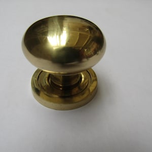 Könnte beinhalten: Ein Türknauf aus massivem Messing mit einer abgerundeten, ovalen Form. Der Knauf sitzt auf einer kreisförmigen Basis mit konzentrischen Ringen. Das Messing hat eine polierte, reflektierende Oberfläche, und das Design ist klassisch und funktional.