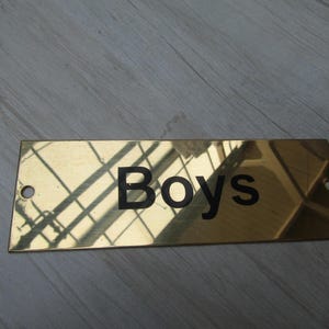 Könnte beinhalten: Ein rechteckiges Messingschild mit einer polierten, reflektierenden goldfarbenen Oberfläche. Das Wort "Boys" ist in der Mitte in fetten, schwarzen, serifenlosen Buchstaben gedruckt. An jeder Seite befindet sich ein kleines Montageloch.