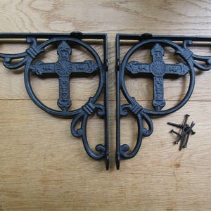 Op de afbeelding: Twee zwarte gietijzeren plankbeugels met een kruisontwerp. Elke beugel heeft een cirkelvormig frame met een sierlijk kruis in het midden. De beugels worden vergezeld door een kleine hoop spijkers, alles tegen een houten achtergrond.