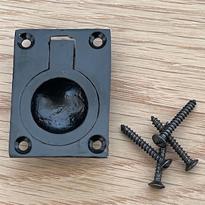 Könnte beinhalten: Ein schwarzer, rechteckiger Einbaugriff mit rundem Griff und vier Schraubenlöchern. Drei schwarze Schrauben befinden sich rechts vom Griff. Die Hardware befindet sich auf einer hellen Holzmaserungsoberfläche.