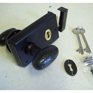 Könnte beinhalten: Ein schwarzes Türschloss-Set mit zwei ovalen Türknöpfen, einer Schlüssellochabdeckung, zwei Schlüsseln und Befestigungsmaterial. Das Schloss hat ein messingfarbenes Schlüsselloch und eine schwarze Metalloberfläche.