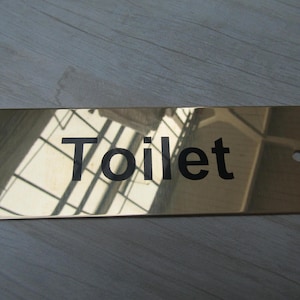 Könnte beinhalten: Ein rechteckiges Schild aus poliertem Messing mit einer reflektierenden goldfarbenen Oberfläche. Das Wort "Toilet" ist mittig in fetten, schwarzen, serifenlosen Buchstaben aufgedruckt. An beiden Seiten befindet sich ein kleines Montageloch.
