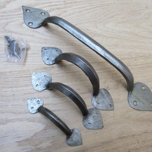 Może przedstawiać: Zestaw czterech metalowych uchwytów do drzwi lub szuflad w stylu antycznym. Każdy uchwyt ma zakrzywiony uchwyt i płyty montażowe w kształcie serca z wstępnie wywierconymi otworami. Dołączona jest również mała torebka ze śrubami.