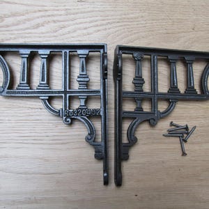 May include: Two black cast iron shelf brackets with a decorative balustrade design. Each bracket features vertical pillars and curved accents. The brackets are accompanied by a small pile of screws.