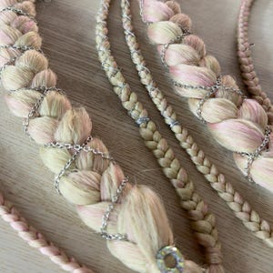 Op de afbeelding: Een set haarextensies met dikke, gevlochten strengen in een mix van lichtblond en zachtroze. De vlechten zijn versierd met fijne zilverkleurige kettinkjes, kleine metalen bedeltjes en een enkele iriserende ronde kraal aan de onderkant.