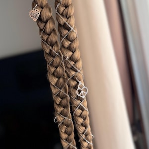 Puede incluir: Dos extensiones de cabello castaño trenzado con detalles plateados. Cada trenza presenta un gran anillo plateado en la parte superior, bandas plateadas envueltas y dijes colgantes. Un dije es un corazón, el otro es un ojo.
