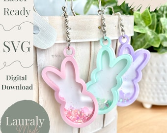 May include: Three pastel-colored bunny-shaped keychains. The keychains are pink, mint green, and lavender, each filled with glitter. The keychains are attached to silver keyrings. The image includes the text "Laser Ready", "SVG", "Digital Download", and "Lauraly Made Designs".