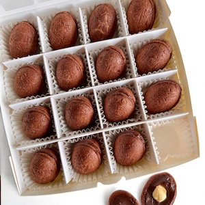May include: A white box containing 12 chocolate truffles, each in a paper cup. Two truffles are outside the box, one cut open to show a hazelnut. The truffles are oval and dark brown in colour.
