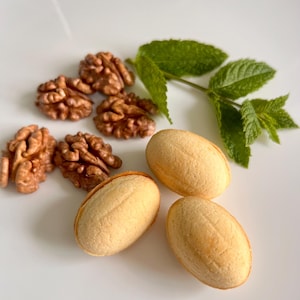 May include: A selection of walnuts, biscuits, and mint leaves presented on a white surface. The walnuts are brown and textured, the biscuits are oval-shaped with a light brown hue, and the mint leaves are green.