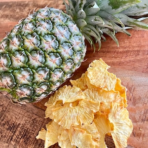 May include: A whole pineapple with green leaves and a pile of dried pineapple slices on a wooden surface.