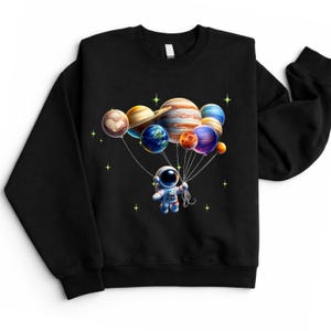 May include: Black sweatshirt featuring a colorful graphic of an astronaut holding planet balloons. The planets include Earth, Jupiter, and Saturn. The design is surrounded by small yellow stars.