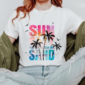 May include: White t-shirt with a colorful graphic that reads "SUN, SEA, SAND" with palm trees and birds. The text is in a gradient of pink, orange, and blue. The shirt is a casual, beach-themed design.