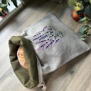 May include: A linen bread bag with a lavender design. The bag is tied with a string and has a green lining. A loaf of bread is inside the bag.
