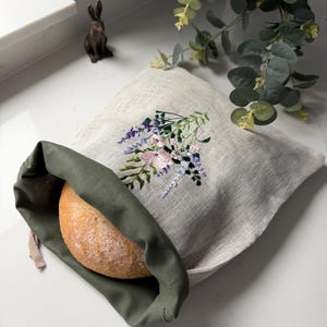 May include: A round loaf of bread sits inside a green linen bread bag. The bag is inside a beige linen bread bag with an embroidered floral design in pink, purple, and green. A small bronze rabbit figurine is in the background.