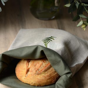 May include: A freshly baked loaf of bread is nestled in a linen bread bag. The bag is light grey with a green fern embroidery. The interior is a darker green. The bread has a golden-brown crust. The bag is designed to keep bread fresh.