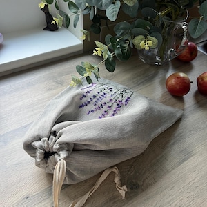 May include: A grey linen drawstring bag with a lavender floral design. The bag is sitting on a wooden surface.