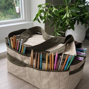 Puede incluir: Un bolso tote de artesanía beige y verde oliva lleno de varias agujas de tejer y ganchillos. El bolso tiene múltiples bolsillos y un asa resistente. Las agujas y los ganchillos son de varios colores y tamaños, lo que sugiere una variedad de proyectos.