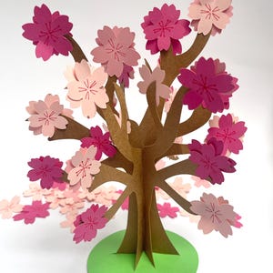May include: A papercraft cherry blossom tree with pink flowers and a brown trunk. The tree is standing on a green base.