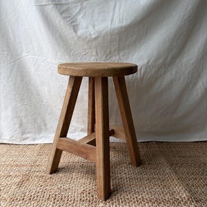 May include: A small, round wooden stool with a natural light brown finish. The stool features a circular seat supported by four straight, angled legs that are connected by horizontal wooden crossbars for stability and structural support.