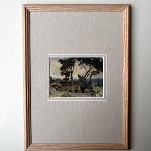 May include: Framed landscape painting featuring a rural scene with trees and buildings. The artwork is set within a light-colored mat and a natural wood frame. The painting's colors include greens, browns, and blues, creating a serene atmosphere.