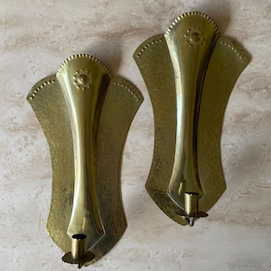 May include: Two vintage brass wall sconces. Each sconce features a fan-shaped backplate with a hammered texture and a central, elongated candle holder. The sconces are made of brass and have a warm, golden hue.