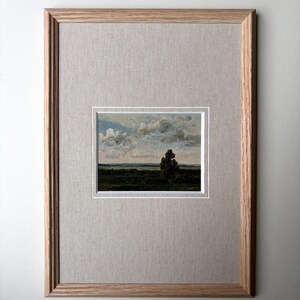 May include: Framed landscape painting featuring a tree in a field under a cloudy sky. The artwork is set within a white mat and a light brown wooden frame, creating a classic aesthetic. The painting's colors include greens, blues, and browns.