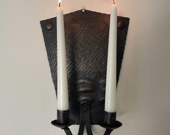 May include: A black metal wall sconce with two white candles. The sconce has a textured, shield-shaped backplate and two candle holders. The candles are lit, with flames at the top. The sconce is mounted on a white wall.