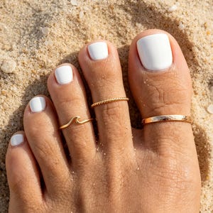 Puede incluir: Primer plano de un pie con esmalte de uñas blanco y tres anillos de oro para los dedos de los pies. Un anillo es una banda simple, otro tiene un diseño de cuerda trenzada y el tercero tiene forma de ola. El pie descansa sobre arena.
