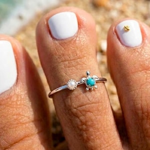 Op de afbeelding: Zilveren teenring met een kleine turquoise schildpad en een witte parel. De ring wordt gedragen op een teen met witte nagellak. De ring is een sieraad met een strandthema.