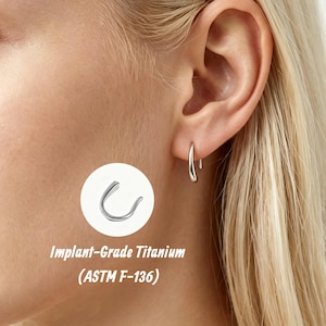 May include: A silver-coloured U-shaped earring made of implant-grade titanium (ASTM F-136). The earring features a smooth, curved design and is shown worn in an earlobe, with a separate view of the jewellery piece displayed in a white circle.