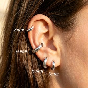 Può includere: Quattro orecchini a cerchio color argento lucido indossati su un orecchio. I cerchi sono etichettati con le misure: 8mm in alto, 12mm al centro, 10mm sul lobo e 8mm sul lobo inferiore. Gli orecchini hanno una finitura metallica liscia e riflettente.