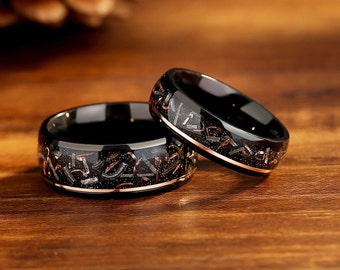 May include: Two black tungsten carbide wedding bands with a rose gold inlay and a pattern of small silver and gold flecks. The bands are on a wooden surface.
