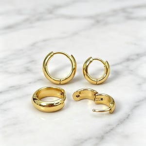 以下が含まれることがあります： 光沢のある滑らかな仕上げのゴールドトーンのフープピアス4点セット。2つは円形で閉じた状態のフープで、残りの2つは厚みのある幅広のフープです。そのうちの1つは半開きになっており、ヒンジのメカニズムが見える状態になっています。