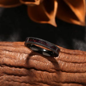 May include: A black ring with a textured band and a dark red, glittery inlay. The ring is set against a blurred background, highlighting the jewelry's design and details. The ring is a piece of jewellery.