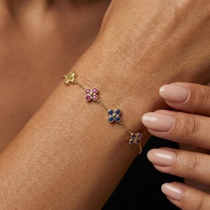 以下が含まれることがあります： 4つの花型のチャームが付いたゴールドのブレスレット。各チャームには、カラフルな宝石のクラスターがセットされています。ブレスレットは手首に着用され、繊細なデザインとジュエリーの鮮やかな色を際立たせています。