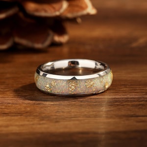 May include: A silver-colored ring with a white opal band and gold flakes. The ring is set against a wooden surface, with a blurred pinecone in the background. The ring's design is simple yet elegant, suitable for everyday wear.