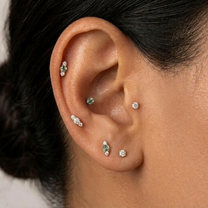 May include: Close-up of an ear adorned with multiple earrings. The earrings include small, round diamond studs, and others with a combination of diamonds and green gemstones. The ear is set against a dark background.