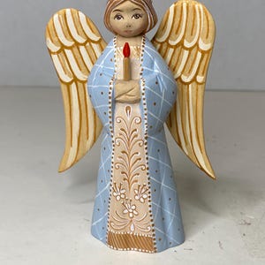 May include: A hand-painted angel figurine with a light blue robe featuring a diamond pattern. The angel holds a candle with a red flame. The wings are beige with white accents. The figurine has a serene expression.