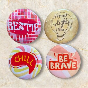 Op de afbeelding: Vier ronde buttons. Linksboven een roze en rood ruitpatroon met de tekst BESTIE. Rechtsboven geel met een illustratie van een gloeilamp en de tekst LET YOUR LIGHT SHINE. Linksonder gele en roze wervelingen met de tekst CHILL. Rechtsonder roze en wit met de tekst BE BRAVE.