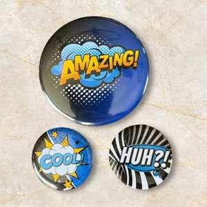 Op de afbeelding: Drie ronde blauwe en witte knoppen met tekst in comic-stijl. De eerste knop zegt "AMAZING!" in gele letters. De tweede knop zegt "COOL!" in gele letters. De derde knop zegt "HUH?!" in gele letters.
