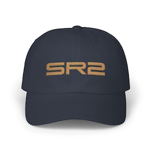 May include: Navy blue baseball cap with the embroidered text "SR2" in a gold color. The cap has a curved brim and a classic six-panel design. The hat is made of a soft, durable material.