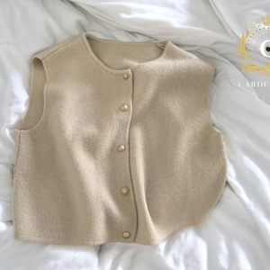 May include: A beige, sleeveless sweater vest with a round neckline and a button-down front. The vest has a soft texture and is buttoned with six small, round buttons. The garment is displayed on a white, textured surface.